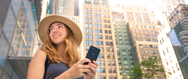 Atractiva mujer positiva sosteniendo el teléfono móvil en la mano mirando la ciudad moderna