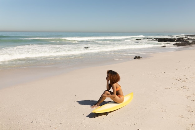 Una atractiva mujer de raza mixta disfrutando del tiempo libre en la playa en un día soleado, divirtiéndose, sentada en su tabla de surf, el sol brillando sobre ella.