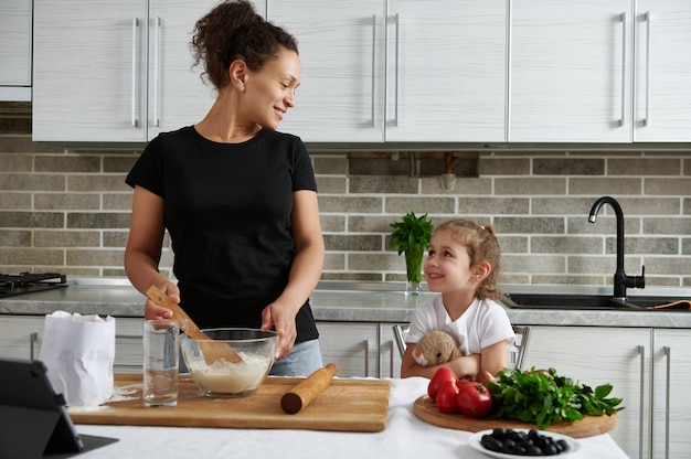 Atractiva mujer de raza mixta, mirando a su linda niña mientras amasa la masa. Madre e hija cocinando juntas en la cocina
