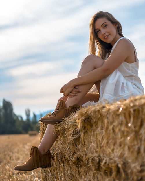 Atractiva mujer rubia con una camisa blanca sentada en un pajar y posando para la cámara