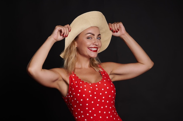 Atractiva mujer rubia caucásica con un traje de baño rojo con lunares blancos y un sombrero de verano de paja sonríe con una hermosa sonrisa con dientes mirando a la cámara posando contra el fondo de la pared negra Copiar espacio