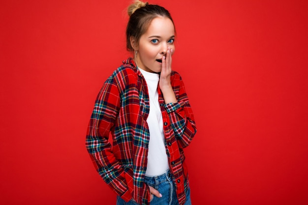 Atractiva mujer rubia joven asombrada que se encuentran aisladas sobre la pared roja vistiendo casual camisa roja y camiseta blanca mirando a la cámara