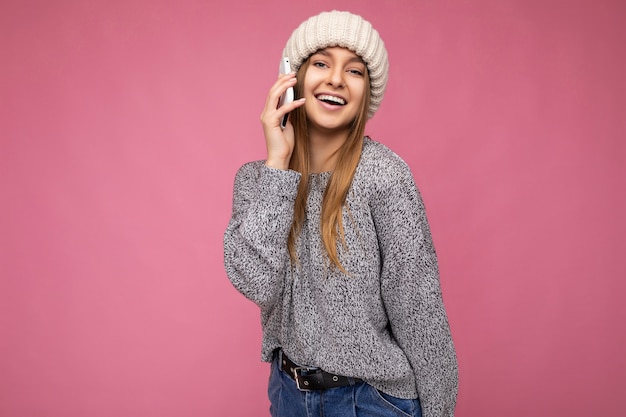 Atractiva mujer rubia joven feliz positiva con suéter gris casual y sombrero beige aislado sobre fondo rosa sosteniendo en la mano y hablando por teléfono móvil mirando a la cámara.