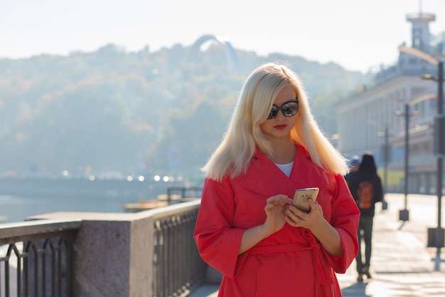 Atractiva mujer rubia madura escribiendo mensaje en el teléfono móvil, posando en los rayos del sol. Copia espacio