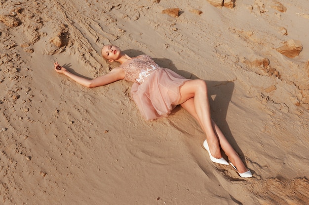 Atractiva mujer rubia posando en vestido bordado en el desierto, acostado sobre arena dorada, a la luz del atardecer.