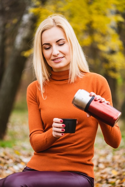 Atractiva mujer rubia en ropa brillante abrir termo de metal y verter té, café en la taza en el fondo de otoño