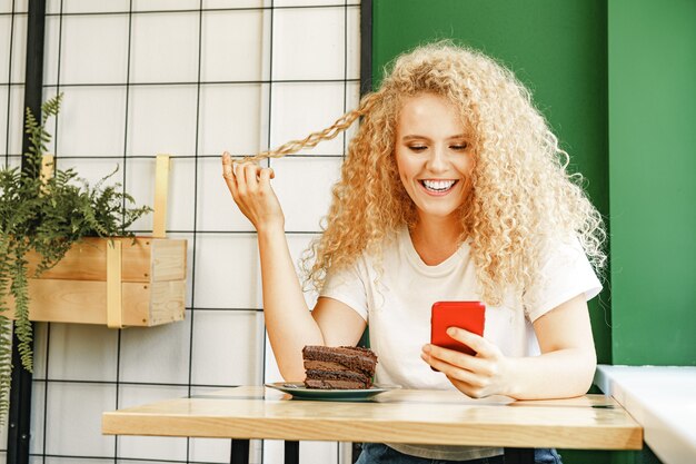 Atractiva mujer rubia sentada junto a la ventana en la cafetería y usando su teléfono móvil