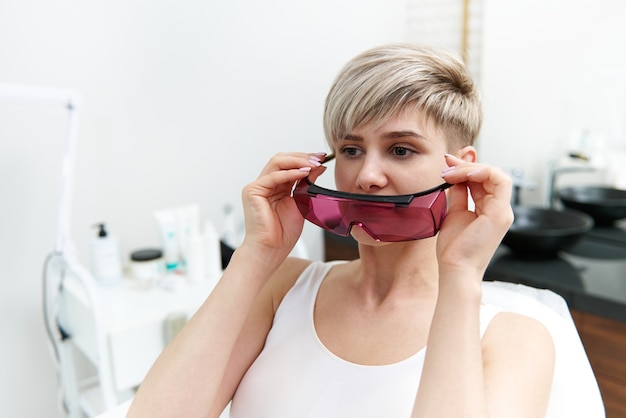 Atractiva mujer rubia sentada en la silla médica en la sala de cosmetología en el salón de spa y ponerse gafas protectoras UV