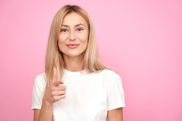 Atractiva mujer rubia sonriendo y eligiéndote apuntando con el dedo a la cámara aislada en el fondo rosa