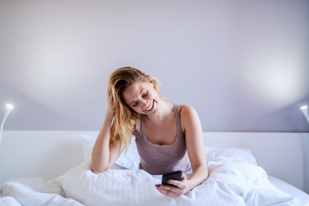 Atractiva mujer rubia sonriente sentada en la cama en el dormitorio por la noche y leyendo el mensaje en el teléfono inteligente