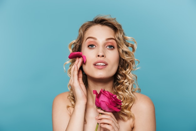 Atractiva mujer semidesnuda con el pelo rizado mirando mientras sostiene una flor rosa aislada sobre una pared azul