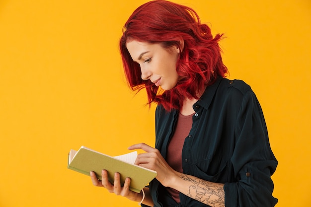 Atractiva mujer seria con pelo rojo rizado sosteniendo y leyendo el libro de ejercicios aislado