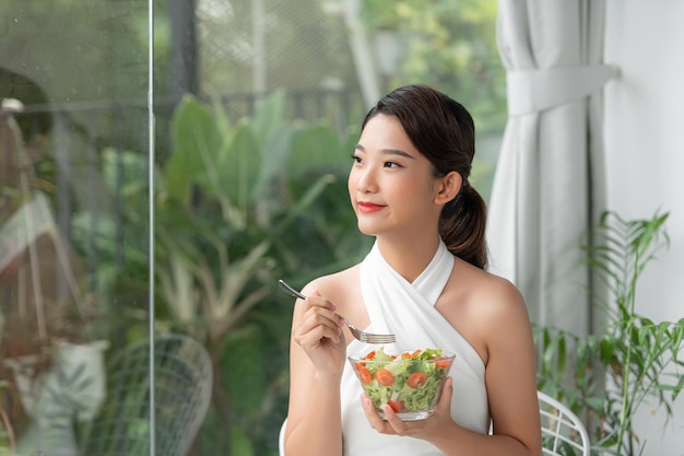 Atractiva mujer sonriente asiática aislada en la sala de estar en casa comiendo ensalada