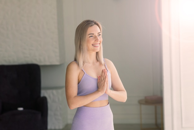 Atractiva mujer sonriente joven practicando yoga, haciendo ejercicio, vistiendo ropa deportiva, sujetador, entrenamiento en casa