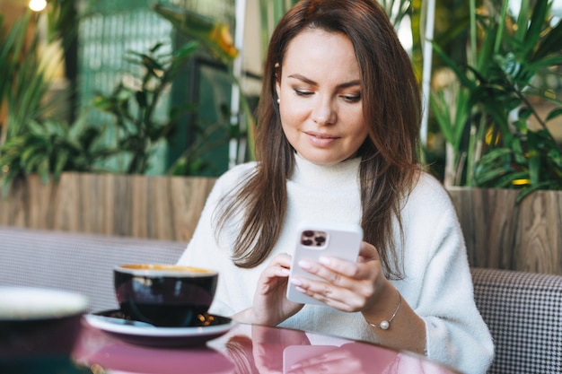 Atractiva mujer sonriente joven en vestido casual blanco con una taza de café usando el teléfono móvil en la cafetería