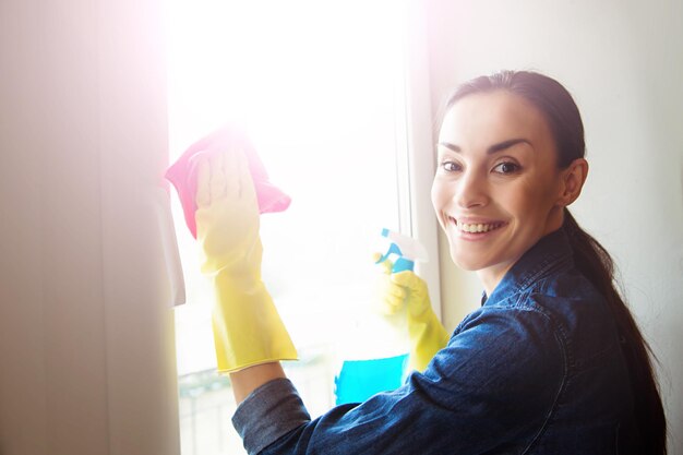 Atractiva mujer sonriente lavando la ventana con spray y trapo