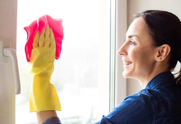 Atractiva mujer sonriente lavando la ventana con spray y trapo