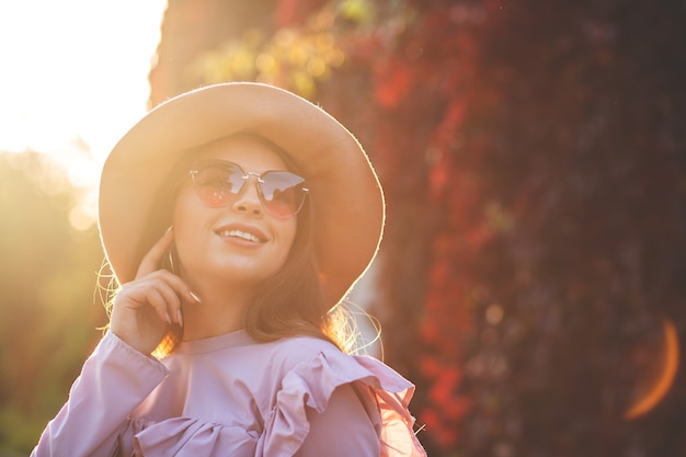 Atractiva mujer sonriente con maquillaje natural con sombrero y gafas de sol, posando en el parque soleado. Espacio para texto