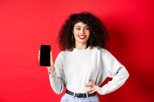 Atractiva mujer sonriente que muestra la pantalla vacía del teléfono inteligente y parece feliz, haciendo publicidad de la tienda o aplicación en línea, de pie contra el fondo rojo.