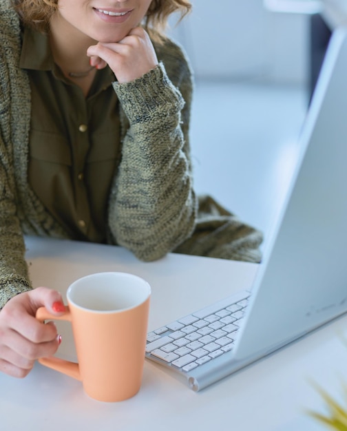 Atractiva mujer sonriente sentada en el escritorio de la oficina sosteniendo una taza de café, se está relajando y mirando hacia otro lado