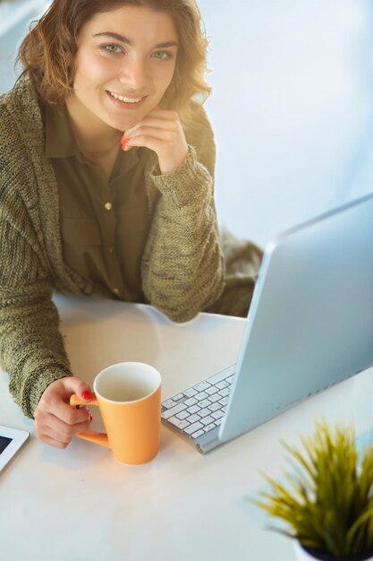 Atractiva mujer sonriente sentada en el escritorio de la oficina sosteniendo una taza de café, se está relajando y mirando hacia otro lado