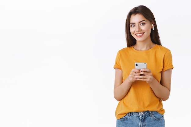 Atractiva mujer tierna moderna con estilo en camiseta amarilla usa auriculares inalámbricos, sosteniendo teléfono inteligente, cámara sonriente complacida y feliz mientras escucha su canción favorita durante el viaje en avión