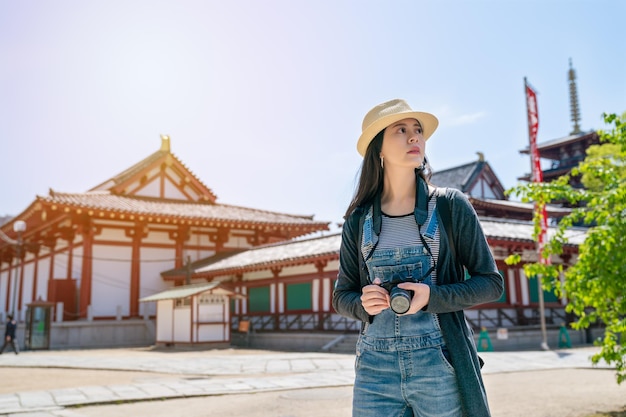 atractiva mujer viajera asiática sosteniendo su cámara y de pie en el hermoso templo de osaka.