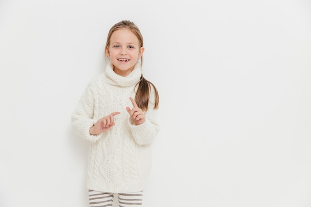 Atractiva niña pequeña en suéter de invierno cálido, escucha una historia positiva de un amigo, se levanta contra blanco