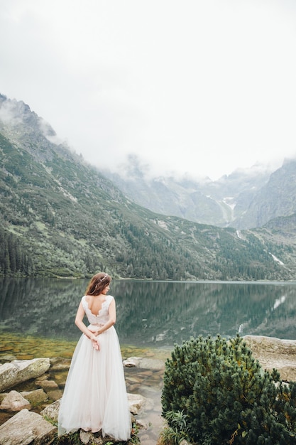 Atractiva novia morena con un hermoso vestido de novia blanco de pie cerca del lago Morskie Oko