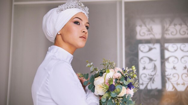 Atractiva novia musulmana con maquillaje profesional en vestido de novia con ramo de flores