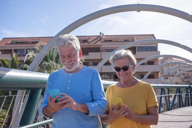 Atractiva pareja de ancianos caminando sobre un puente en la ciudad Mirando su propio teléfono móvil y sonriendo Ancianos modernos y tecnológicos