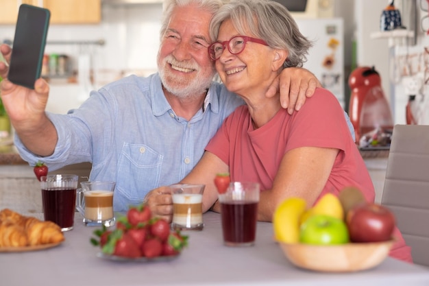 Atractiva pareja de ancianos despreocupada sonriendo en una videollamada con un teléfono inteligente mientras desayunan en casa Personas mayores relajadas y felices disfrutando de una alimentación saludable