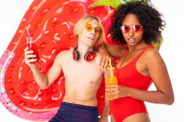 Atractiva pareja chico europeo y chica africana en trajes de baño con gafas de sol y auriculares con cócteles en sus manos