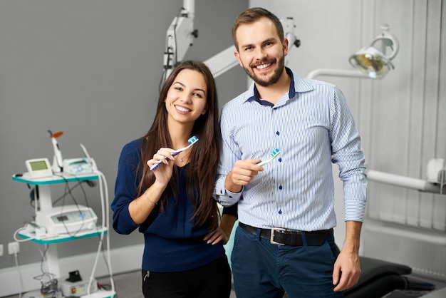 Atractiva pareja está de pie en el consultorio dental del equipo y sosteniendo cepillos de dientes con pasta. Los jóvenes tienen una sonrisa blanca como la nieve y miran a la cámara