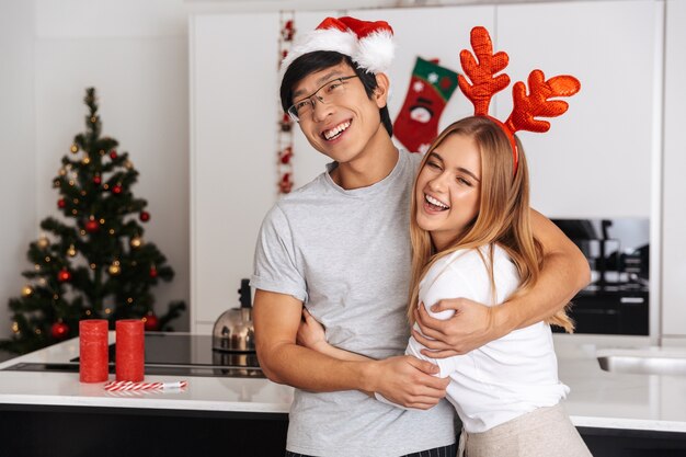 Atractiva pareja, hombre y mujer vestidos con traje de Navidad, de pie en la cocina luminosa y abrazándose juntos