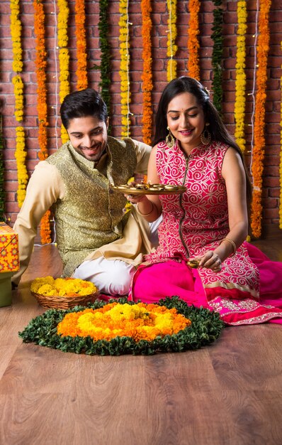 Atractiva pareja india en ropa tradicional celebrando el Festival de Diwali, cumpleaños o aniversario con regalos sorpresa y dulce laddoo sobre fondo decorado con flores de caléndula