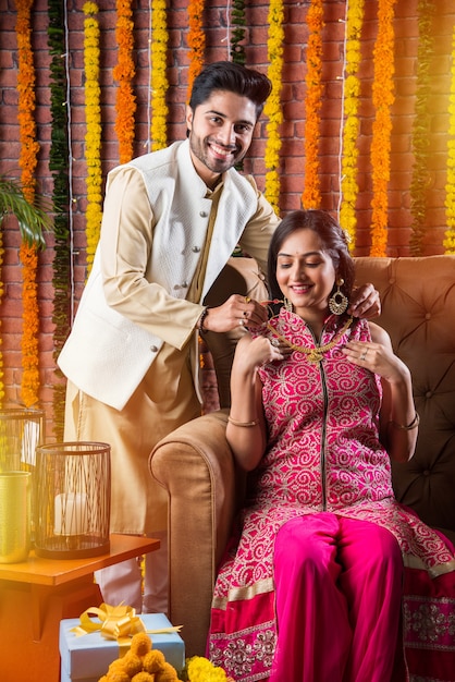 Atractiva pareja india en ropa tradicional celebrando el Festival de Diwali, cumpleaños o aniversario con regalos sorpresa y dulce laddoo sobre fondo decorado con flores de caléndula