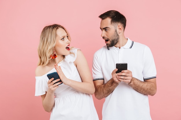 Atractiva pareja joven curiosa de pie juntos aislados sobre rosa, mediante teléfono móvil