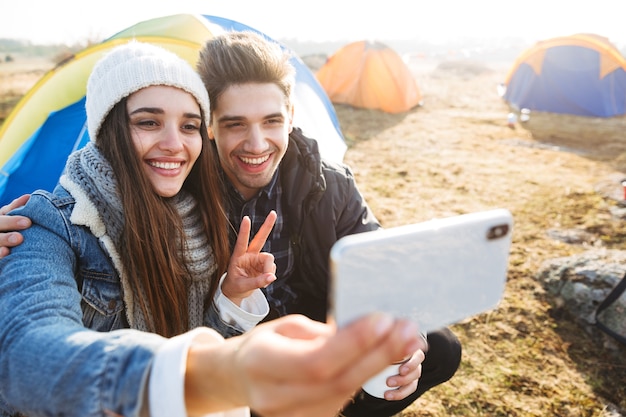 Atractiva pareja joven descansando mientras está sentado en la carpa al aire libre, sosteniendo una taza y un termo, tomando un selfie