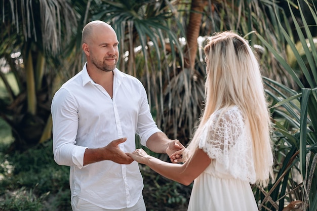 Atractiva pareja joven de moda en ropa blanca cogidos de la mano y posando juntos con el telón de fondo de palmeras. Hermosa rubia de pelo largo y un hombre calvo están de vacaciones. Phuket, Tailandia