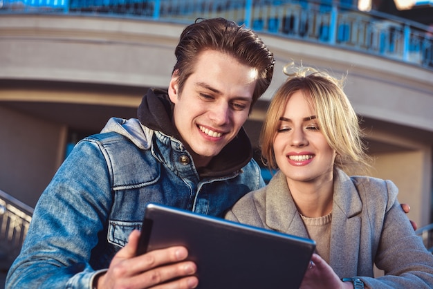 Atractiva pareja joven sentada en el suelo en la calle urbana leyendo información en una tableta con una sonrisa.