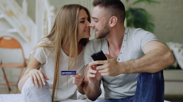 Atractiva pareja joven con teléfono inteligente y compras con tarjeta de crédito en Internet sentada en la cama en casa