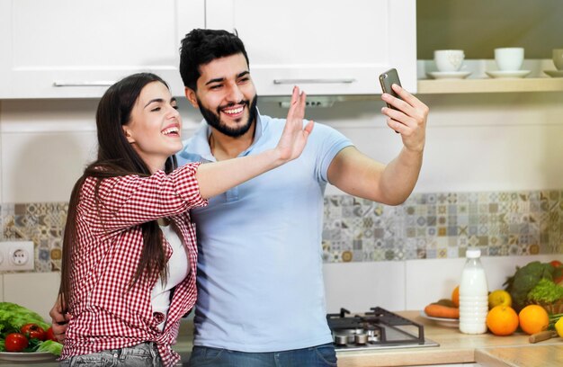 Atractiva pareja joven tomándose selfie con un smartphone usando ropa casual y elegante divirtiéndose juntos en una cocina acogedora