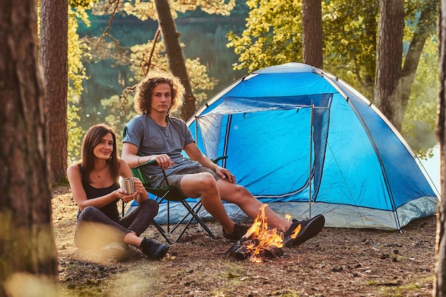 Atractiva pareja de jóvenes relajándose cerca de la hoguera en un bosque verde y soleado. Tienen carpa azul.