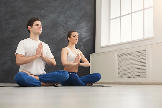 Atractiva pareja practicando yoga sentado en padmasana. Hombre y mujer joven en posición de loto haciendo ejercicio de respiración en la colchoneta en el interior del club deportivo, espacio de copia