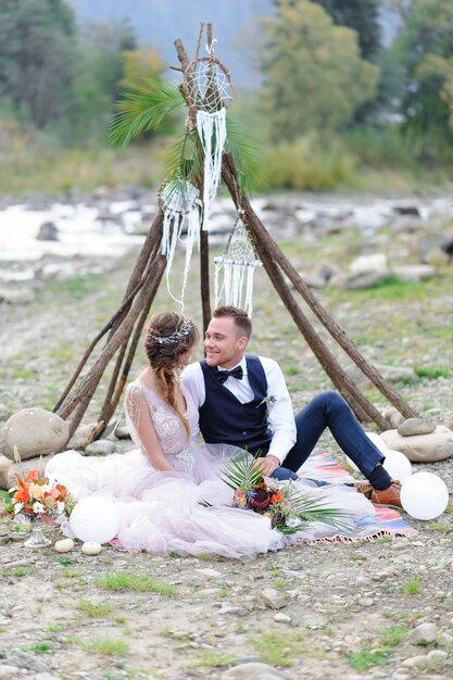 Una atractiva pareja de recién casados, un momento feliz y alegre. Hombre y mujer abrazan y besan en ropa de vacaciones. Boda Cermonia con decoración al estilo Boho junto al río al aire libre.