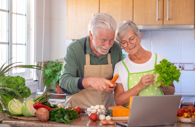 Atractiva pareja senior feliz preparando verduras juntos en la cocina de la casa buscando menú en la computadora portátil Personas mayores caucásicas disfrutando de una alimentación saludable