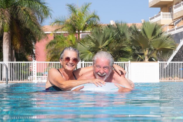 Atractiva pareja senior flotando en la piscina jugando con colchón inflable. Felices jubilados disfrutando del sol y las vacaciones de verano