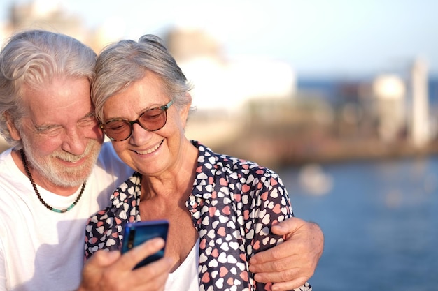 Atractiva pareja senior sonriendo al aire libre en el mar mediante teléfono móvil. Pareja caucásica de pelo blanco disfrutando de la jubilación y la luz del atardecer