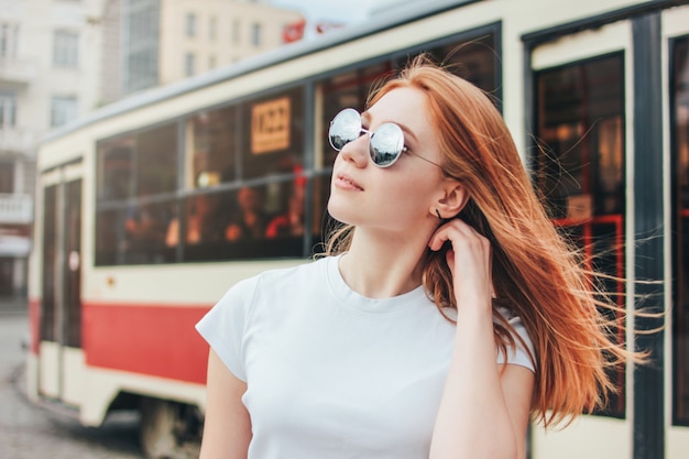 Atractiva pelirroja niña sonriente en gafas de sol en ropa casual sobre fondo de tranvía en la calle